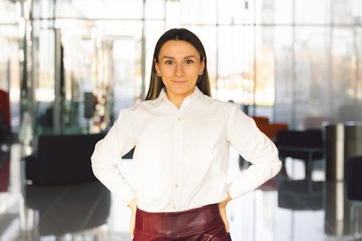 Portrait of a smiling woman in white blouse standing confidently in a bright, modern office space.